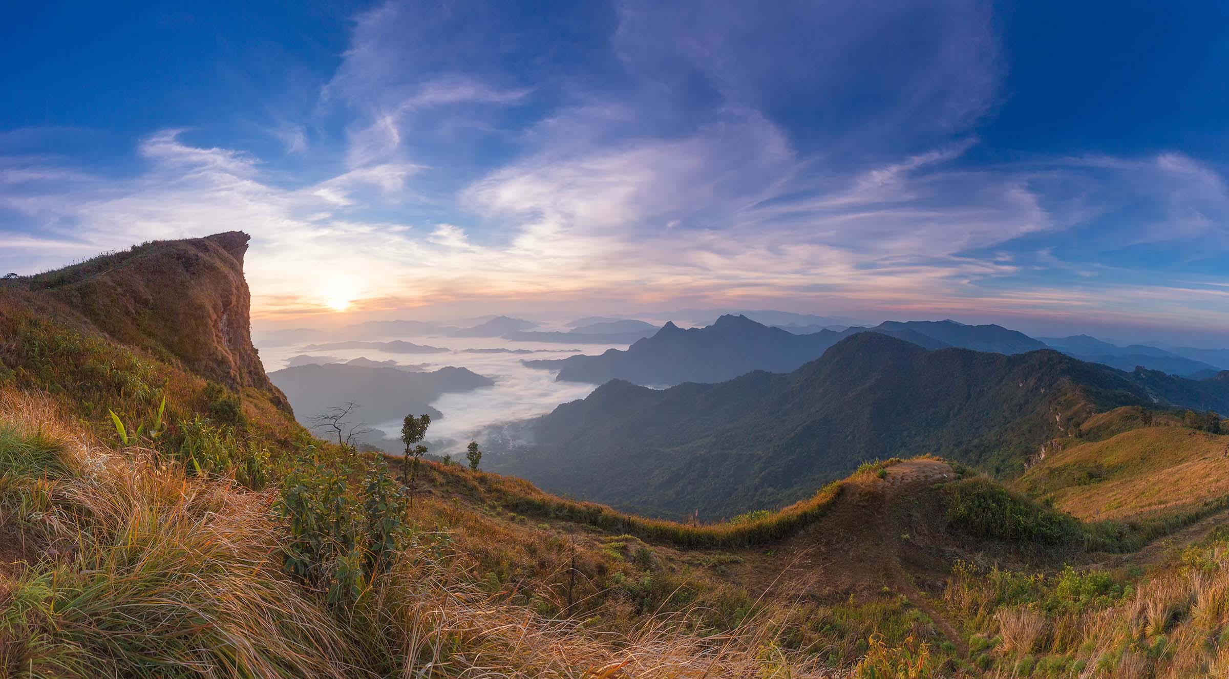 A stunning sunrise over a mist-filled valley, viewed from a mountain ridge with grassy slopes and distant rugged mountains. The sky glows in hues of blue, pink, and orange, casting warmth over the landscape.